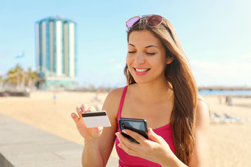 Excited young woman buying online with smart phone reading credit card number outdoor on summer