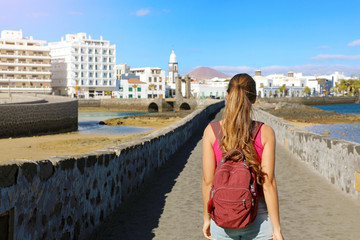 Rear view of traveler girl carrying backpack on her back. Young tourist woman visiting Arrecife...