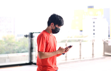 A young man are using a smartphone wearing a black mask on the face for protection against dust pm2.5 and viruses from the air in BTS station in Bangkok, Thailand