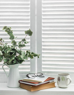 Stack of books,  notebook, glasses,  jug of dry eucalyptus branches on a light window with wooden rustic blinds. Cozy home