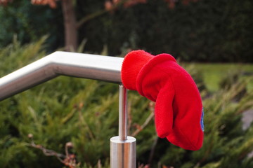A red child's little glove or mitten is lost and lies on a shiny metal railing against a background of green plants. Lost and found