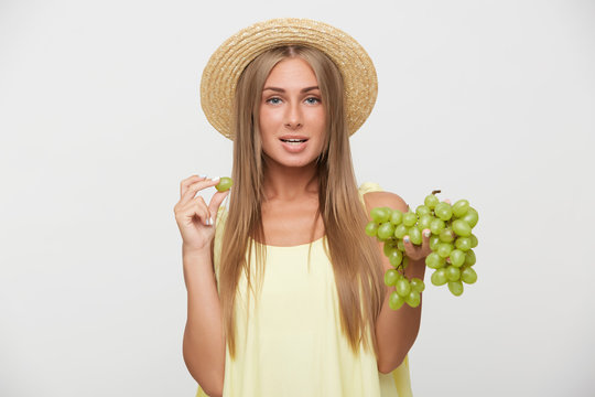 Studio photo of pretty young blue-eyed blonde female with natural makeup keeping green grapes in raised hands while looking at camera with calm face, standing over white background