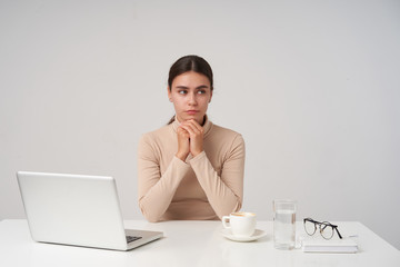 Puzzled young brunette attractive business lady folding hands under her chin while looking thoughtfully aside, dressed in formal clothes while posing over white background