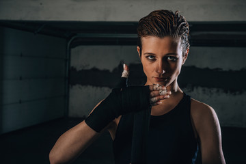 Picture of focused young woman preparing her black bandages for boxing inside a garage.
