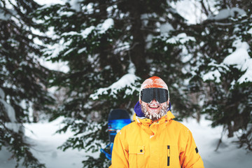 Cheerful smiling snowboarder with snow on his beard and hat stay in yellow jacket in the winter forest on powder day.