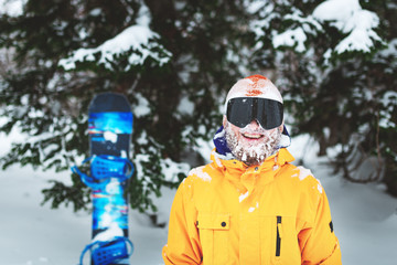 Close up portrait of bearded snowboarder or skier wearing black goggles and orange hat with snow on face standing in mountain forest and smiling. Happy people vacation.