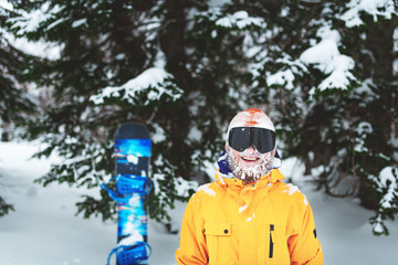 Fototapeta na wymiar Cheerful smiling snowboarder with snow on his beard and hat stay in yellow jacket in the winter forest on powder day.