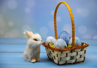 A fluffy toy Easter Bunny sits by a basket of blue colored eggs.