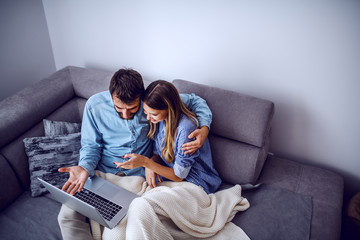 Top view of cheerful cute caucasian couple sitting on sofa covered with blanket and using laptop for internet surf. Living room interior.