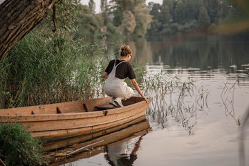 Fototapeta na wymiar Young woman in a beige overalls stands in wooden fishing boat. Sunset over the river in autumn. Romantic view of boat in the pond and forest.