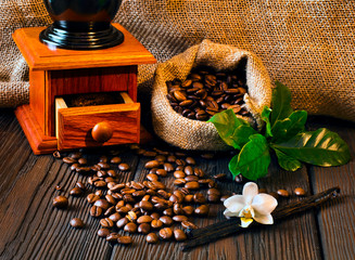 Coffee beans in jute bag with coffee grinder and vanilla sticks on a wooden table.