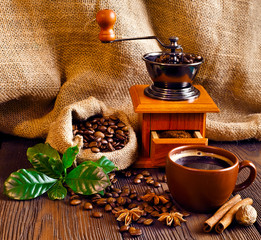 Coffee beans in jute bag with coffee grinder, spices, green leaves and coffee cup on a wooden table.