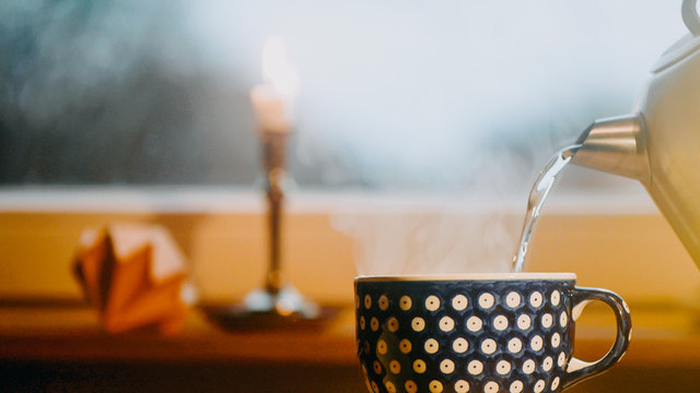 hei&szlig;es Wasser wird aus Wasserkocher in den Becher f&uuml;r Tee gegossen. Das Wasser Dampft. Im Hintergrund steht eine brennende Kerze. Geringe Sch&auml;rfentiefe