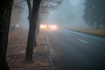 Heavy fog on the road in the early morning. City street with cars in the distance is in poor...