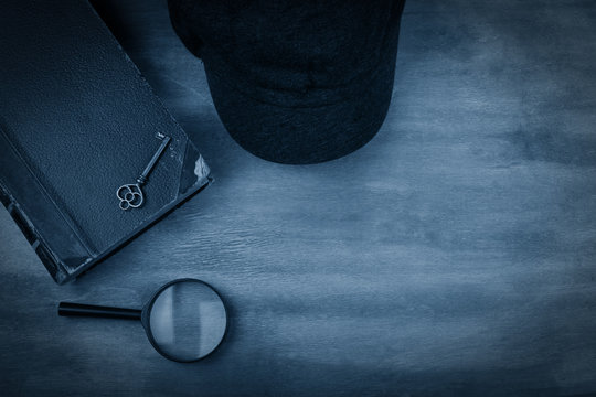 Old book, a key, a detective cap and a magnifying glass against a worn wooden background with dark edges toned in the classic blue color (top view) as the crime story concept