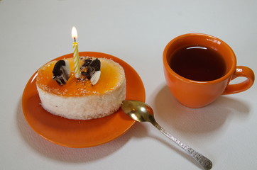  On a white background, an orange ceramic cup with tea and a saucer with cake and a lit candle.