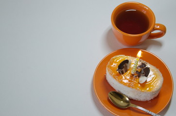  On a white background, an orange ceramic cup with tea and a saucer with cake and a lit candle.