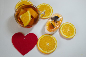 On a white background is a glass cup of tea with orange slices, a light, low-calorie Exotica cake and a red decorative heart.