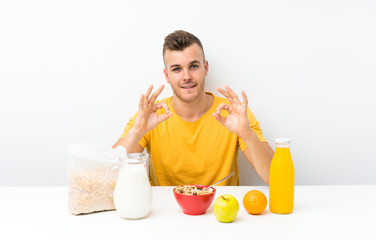 Young blonde man having breakfast showing an ok sign with fingers