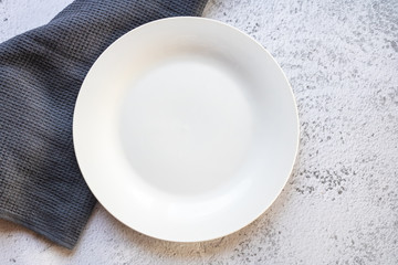 Empty white plate next to a gray kitchen towel on white concrete