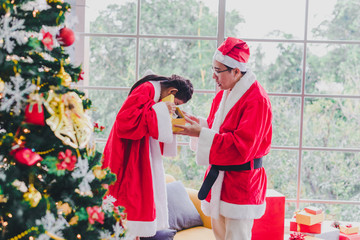 Fototapeta premium Asian father and daughter wear Santa Claus outfits. He gave her gifts on the occasion of Christmas and New Year. In the living room decorated the room to celebrate the important day of the family.