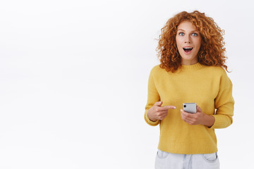 Astonished, excited lucky ginger girl with curly natural hairstyle, hold smartphone, pointing...