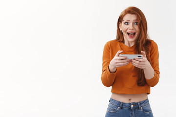 Excited, amused good-looking redhead female in cropped sweater, scream astonished as winning mobile game, passed hard level in online shooter, holding smartphone horizontally, look surprised