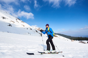 Fototapeta na wymiar Handsome tourist in colorful clothing and sunglasses with backpack trekking on skis in deep snow on background of bright blue sky and beautiful mountain. Winter vacations, active lifestyle concept.