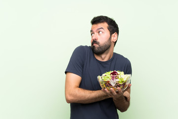 Young handsome man with salad over isolated green wall making doubts gesture while lifting the shoulders