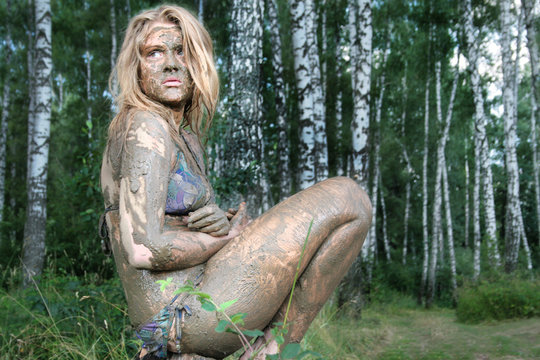 A girl with a mud mask sits in a forest at an ecological resort. Beautiful blonde - dirty, wild and alone in the forest