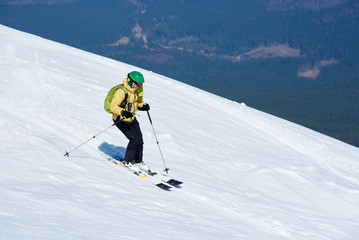 Professional skier in helmet with backpack riding skis fast down steep snowy mountain slope on background of deep white snow and green spruce trees. Winter holidays, sport and recreation concept.