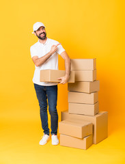 Full-length shot of delivery man among boxes over isolated yellow background pointing to the side...