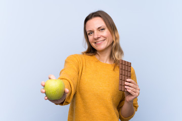 Blonde woman over isolated blue background taking a chocolate tablet in one hand and an apple in...