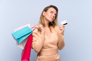 Blonde woman over isolated blue background holding shopping bags and a credit card and thinking