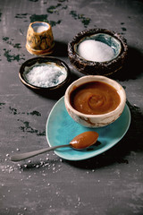 Obraz na płótnie Canvas Homemade salted caramel sauce with grains of fleur de sel salt. Ingredients in ceramic bowls above. Grey textured canvas background.