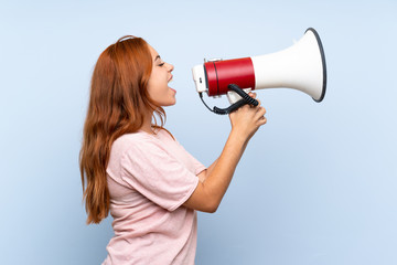Teenager redhead girl over isolated blue background shouting through a megaphone