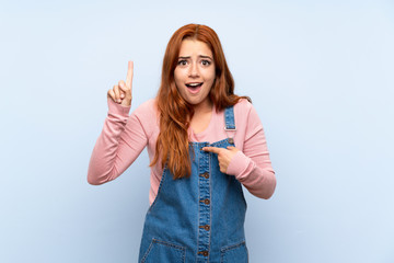 Teenager redhead girl with overalls over isolated blue background with surprise facial expression