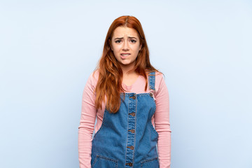 Teenager redhead girl with overalls over isolated blue background having doubts and with confuse...