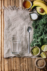 Glass for green healthy vegan smoothie, glass straw. Ingredients above. Kale, bananas, avocado, lime, non-diary milk, honey, matcha powder and seeds over wooden plank cloth background. Flat lay, space