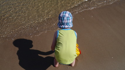 Dziecko na plaży bawi się w piasku nad morzem. Chłopiec z wiaderkiem i łopatką buduje zamki z piasku. Ma założoną czapeczkę przeciwsłoneczną. Dobrze się bawi. - obrazy, fototapety, plakaty