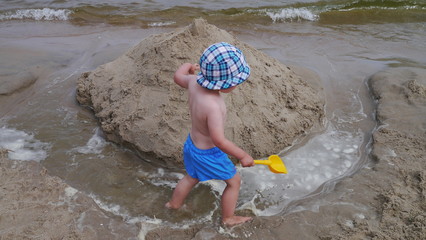 Dziecko na plaży bawi się w piasku nad morzem. Chłopiec z wiaderkiem i łopatką buduje zamki z piasku. Ma założoną czapeczkę przeciwsłoneczną. Dobrze się bawi.