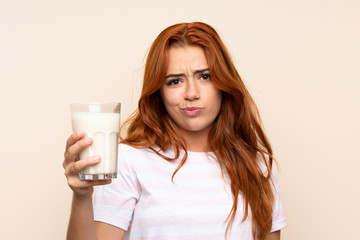 Teenager redhead girl holding a glass of milk over isolated background with sad expression