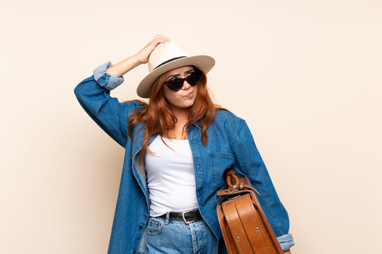 Redhead traveler girl with suitcase over isolated background having doubts and with confuse face expression
