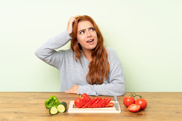 Teenager redhead girl with vegetables in a table having doubts and with confuse face expression