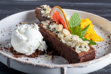 Brownie chocolate dessert with a scoop of white ice cream, slices of mango and a slice of grapefruit. In the background is a cappuccino. On a dark background.