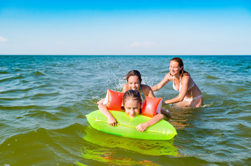 Obraz premium Happy mother and two small positive daughters bathe and swim in the sea with an air mattress on a sunny summer day. Vacation concept with kids. Copyspace