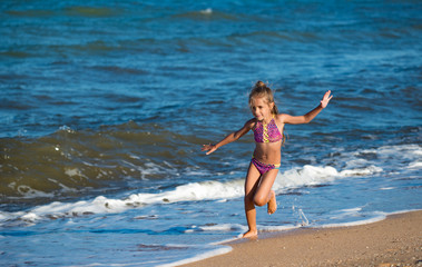 Happy little girl runs along the sandy shore near the calm sea waves on a sunny warm summer day. Cool summer family vacation concept.