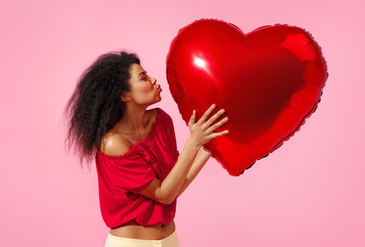 Happy girl holds red heart shape balloon. Photo of smiling young girl in love on pink background. Valentine's Day