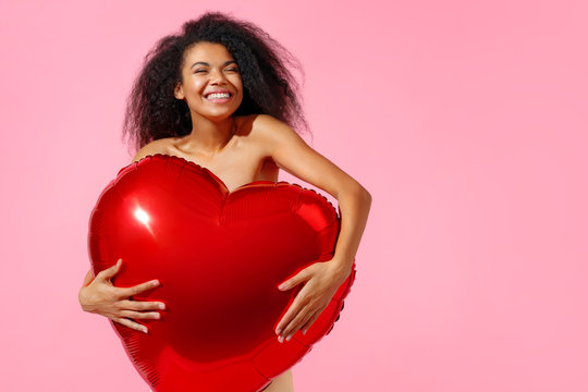Happy girl holds red heart shape balloon. Photo of smiling young girl in love on pink background. Valentine's Day