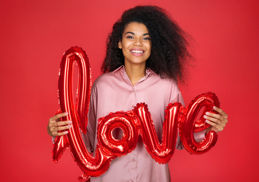 Happy girl holds letter shaped balloon. Photo of smiling young girl in love on red background. Valentine's Day